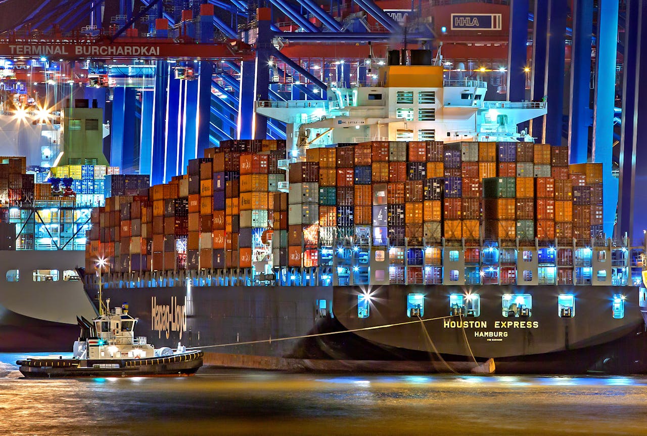 hero-img-01 A brightly lit cargo ship at Hamburg harbor with stacked containers and a tugboat.
